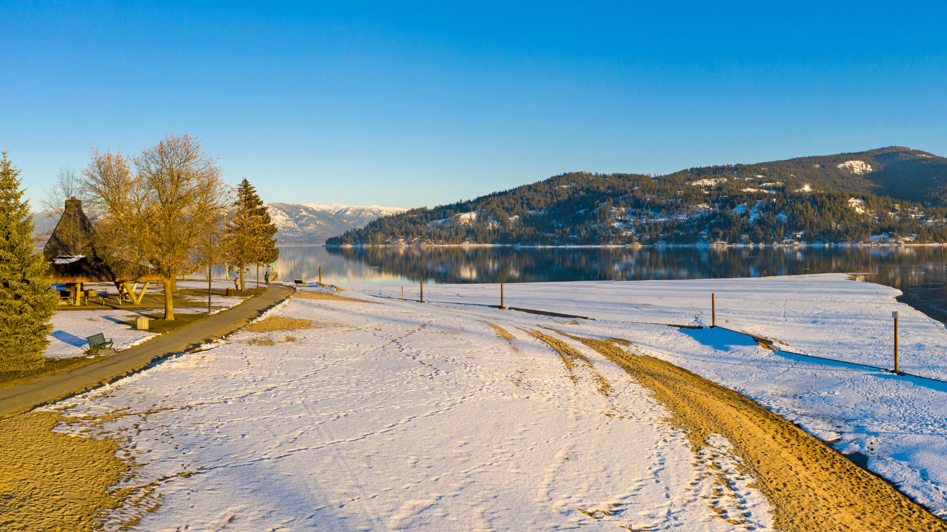 An aerial view of a snow-covered sandy beach in a park setting, where the snow meets the calm, reflective waters of a large lake surrounded by forested mountains under a bright blue sky.