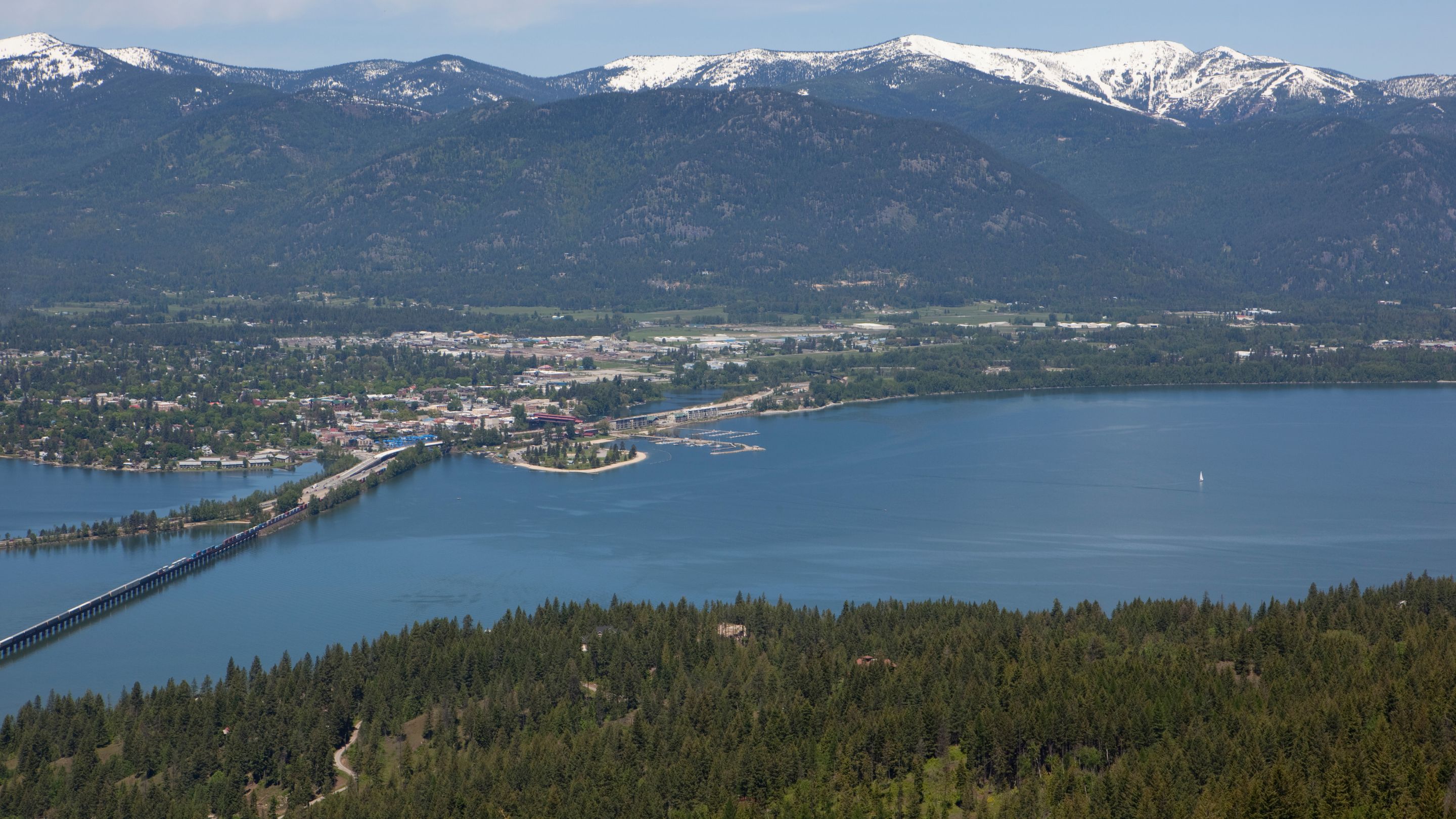 An aerial panoramic view of the town of Sandpoint, Idaho, situated on the blue waters of Lake Pend Oreille, with a long bridge extending across the water and snow-capped mountains visible in the distance.