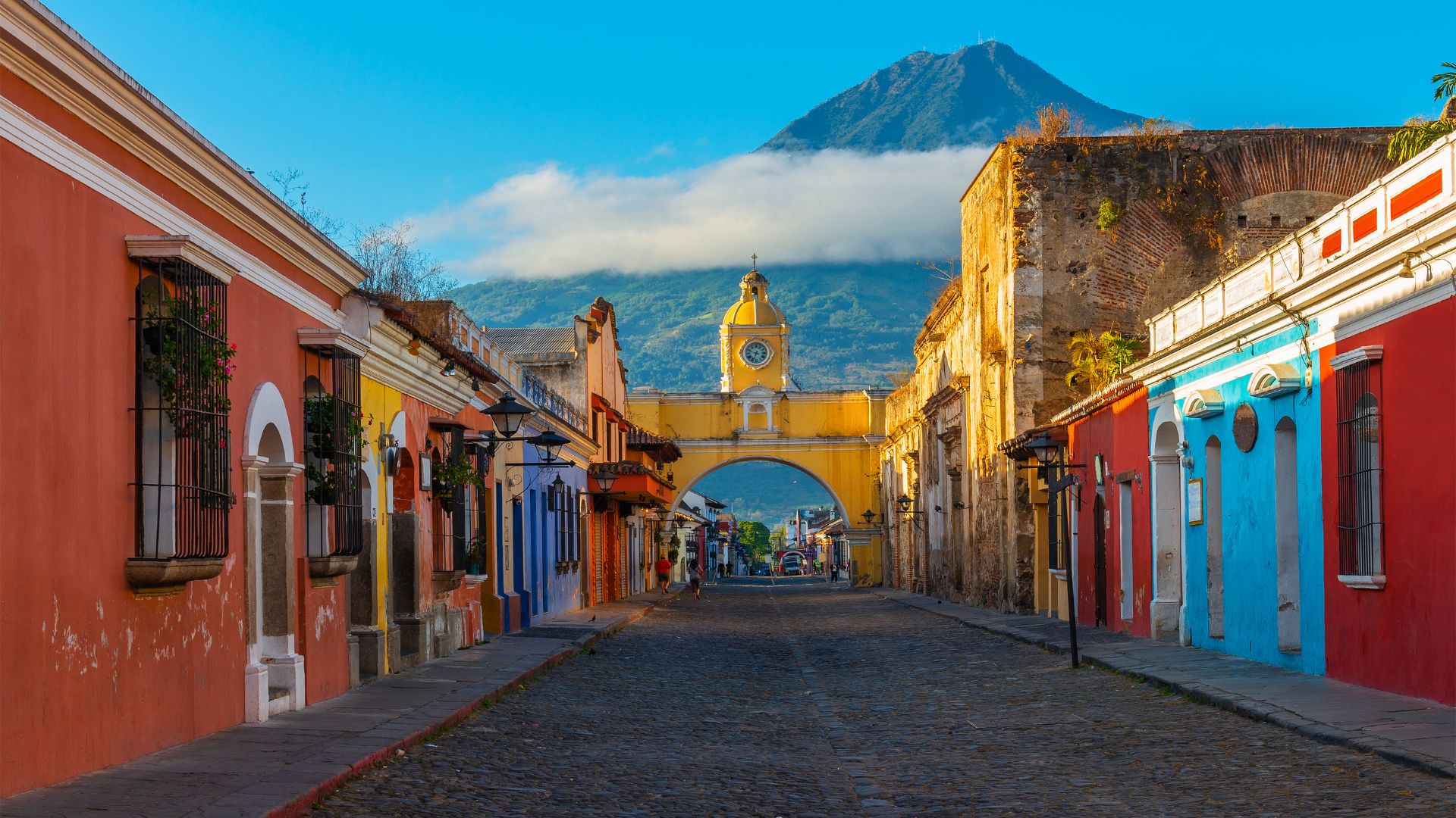 The yellow Santa Catalina Arch spanning a cobblestone street in Antigua, Guatemala, with colorful buildings and a large, cloud-ringed volcano in the background.
