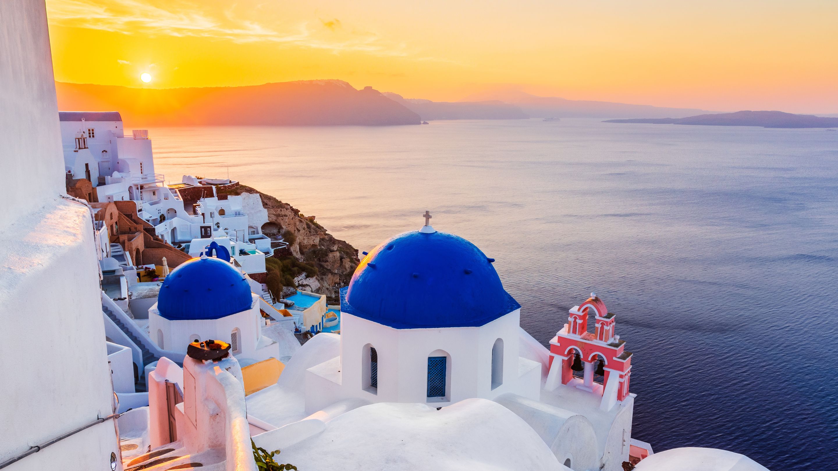 A panoramic sunset view of the iconic, cliffside village of Oia in Santorini, with traditional white buildings, vibrant blue-domed churches, and a small pink bell tower overlooking the deep blue Aegean Sea and distant volcanic mountains.