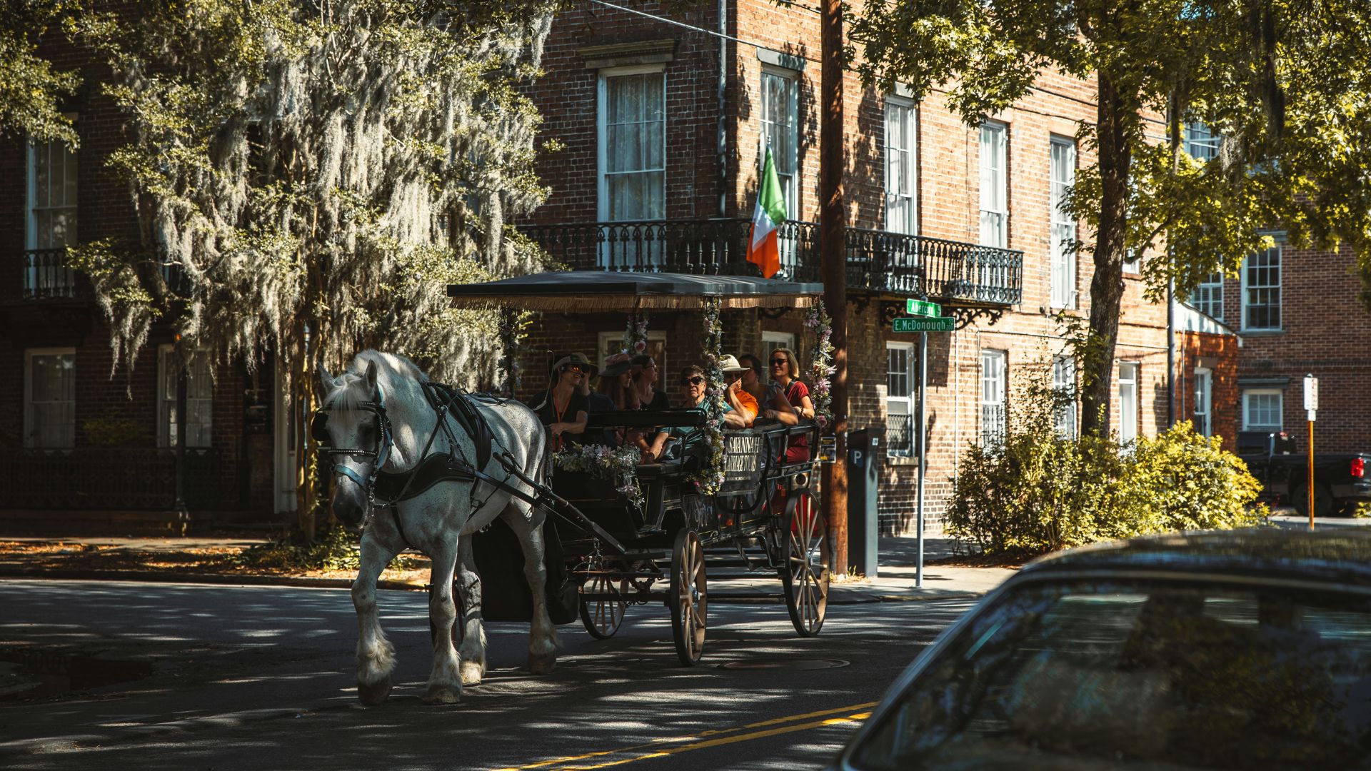 A horse-drawn carriage carrying passengers moves down a sunny, historic brick-lined street in Savannah, Georgia, with a large live oak tree draped in Spanish moss and colonial-style buildings in the background.
