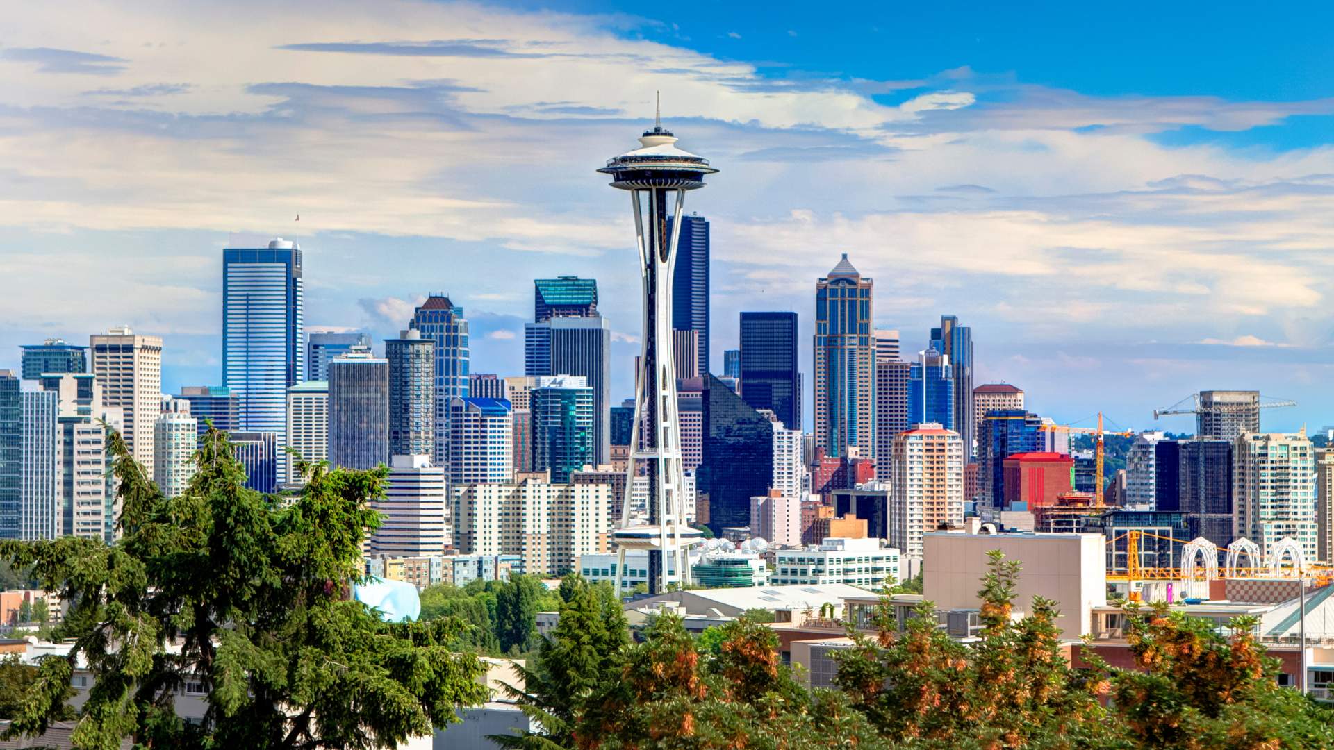 A panoramic daytime view of the Seattle, Washington, skyline featuring the iconic Space Needle and numerous high-rise buildings set against a partially cloudy blue sky, with lush green trees in the foreground.