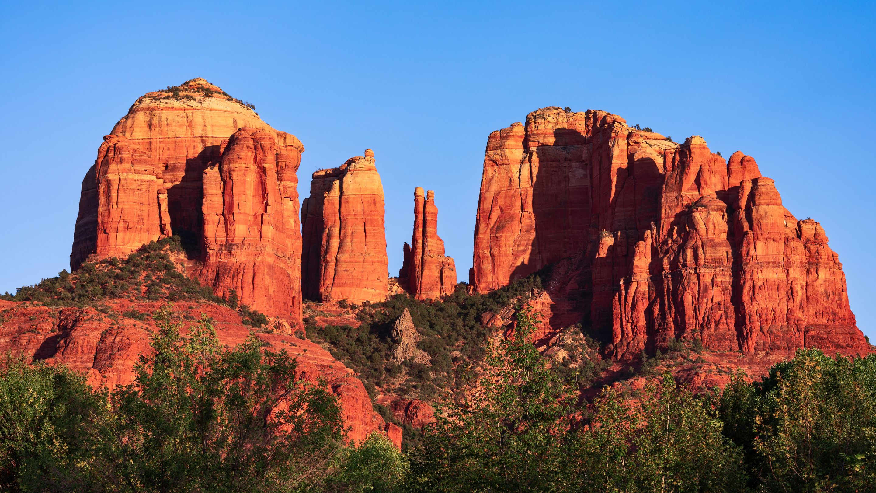 A wide view of the towering red sandstone spires of Cathedral Rock in Sedona, Arizona, against a clear blue sky, with dense green vegetation in the foreground.