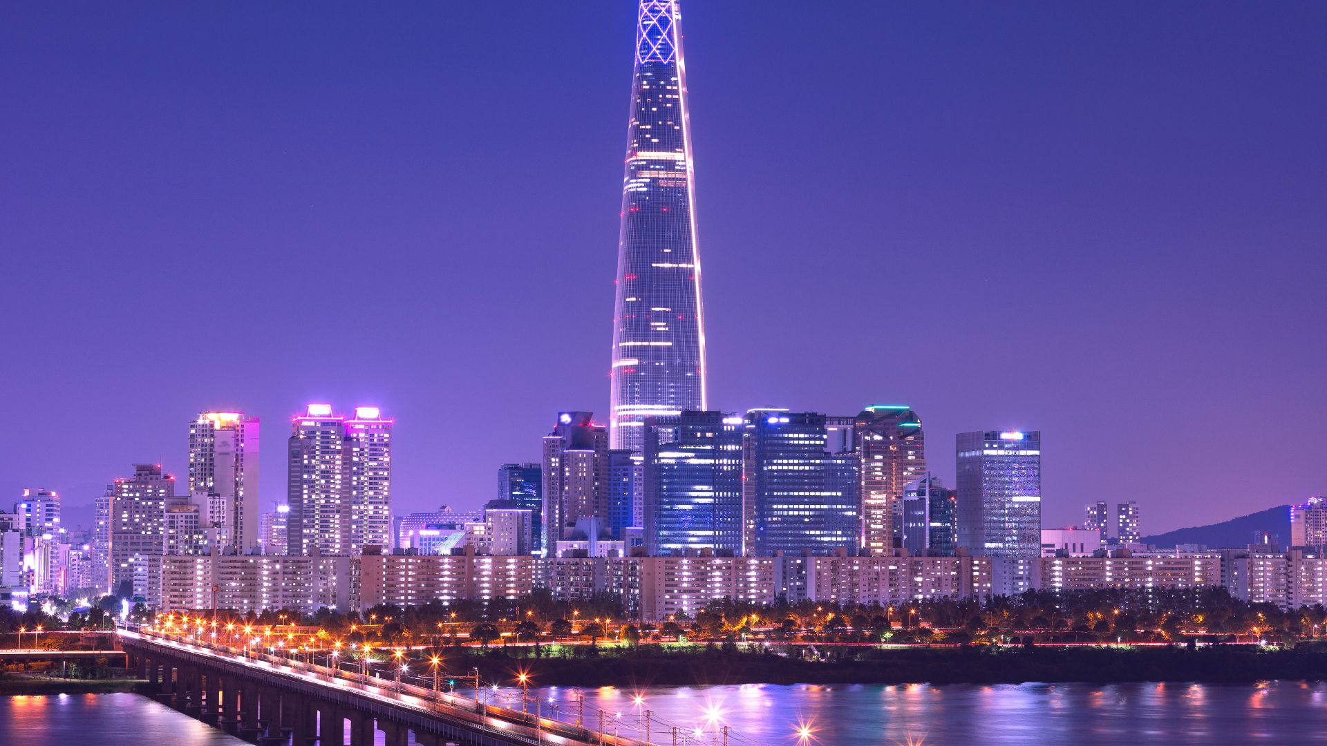 A nighttime panoramic cityscape view of Seoul, South Korea, with the iconic, spired Lotte World Tower standing prominently amidst other illuminated skyscrapers across the dark water of the Han River.