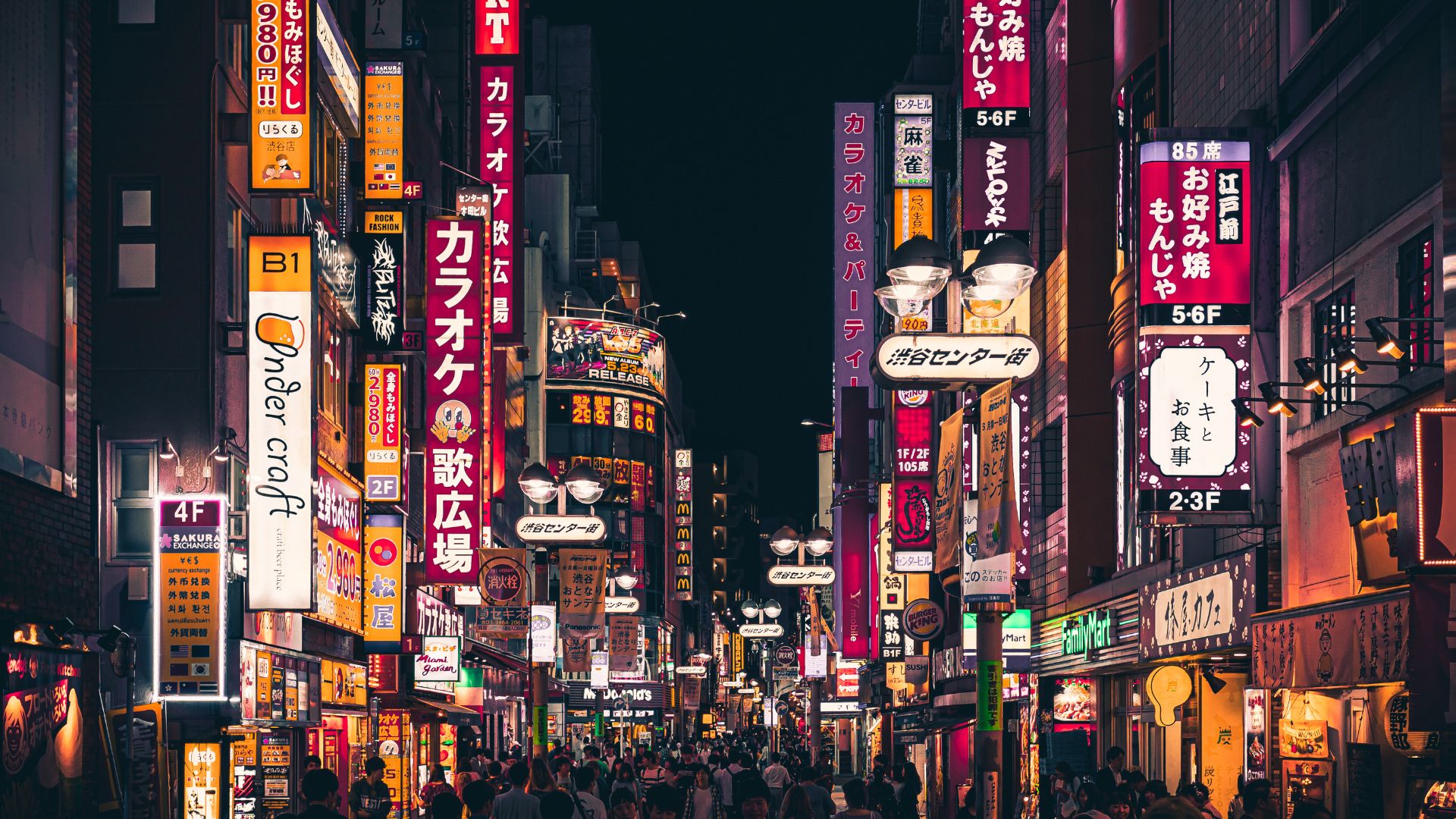 A vibrant, nighttime street scene in Shibuya, Tokyo, featuring narrow streets crowded with people and lined with tall buildings covered in bright, colorful Japanese neon signs.