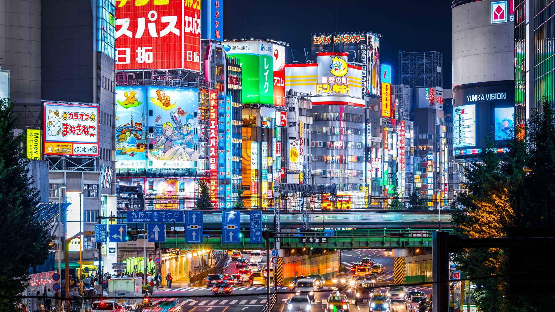 A vibrant nighttime photograph of a busy street in Tokyo's Shinjuku district, densely packed with brightly lit commercial buildings, numerous colorful neon signs, and a busy street intersection with vehicle traffic.