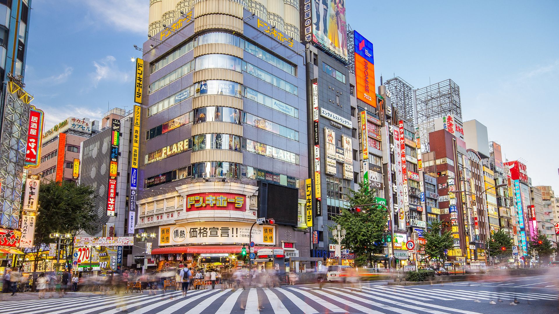 A busy daytime street intersection in Shinjuku, Tokyo, with a multi-story, rounded corner building and a white crosswalk filled with people crossing the road.