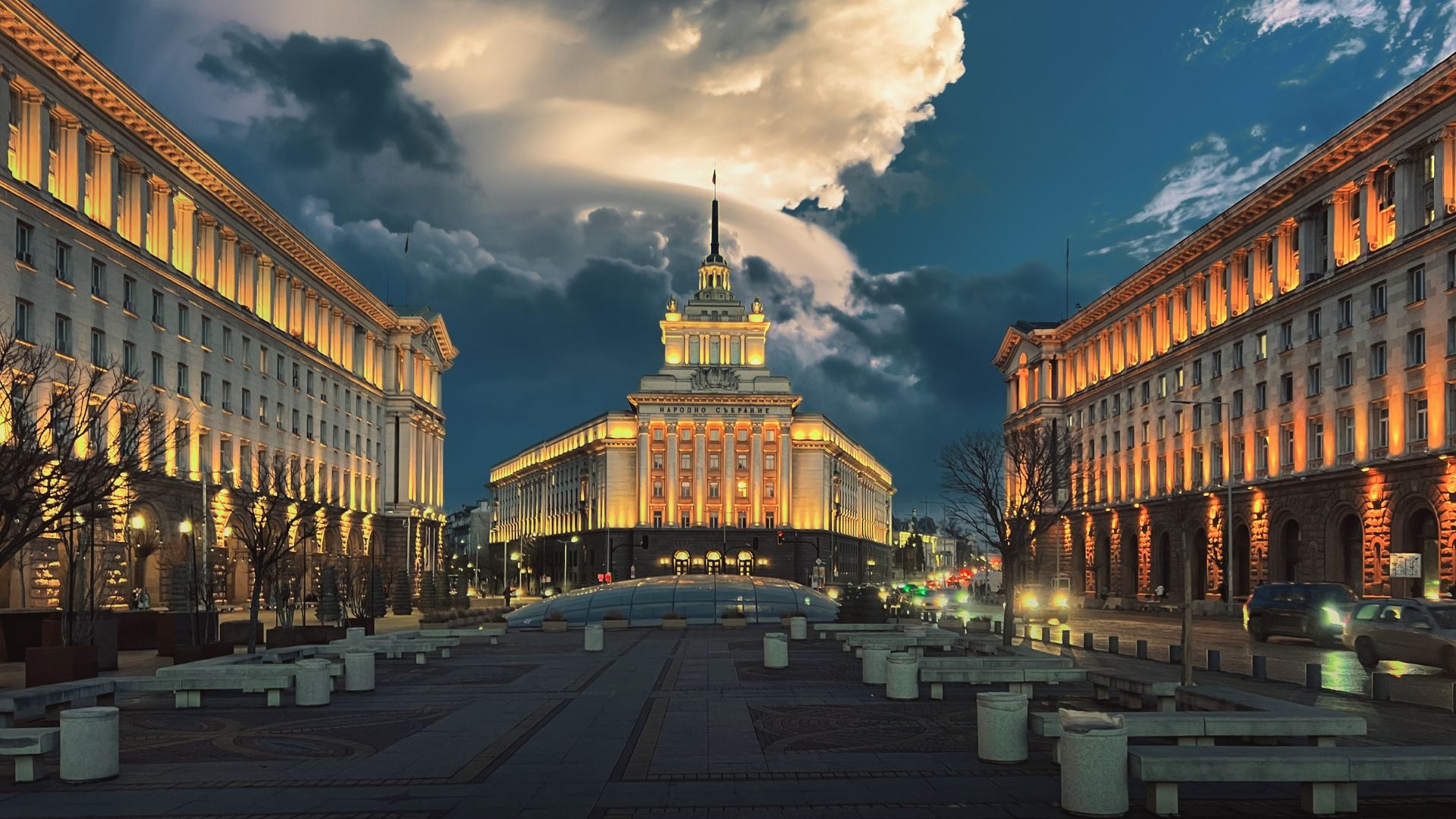 A dramatic, twilight view of the illuminated government buildings forming the Largo architectural complex in central Sofia, Bulgaria, set against a dark blue sky with large white clouds.