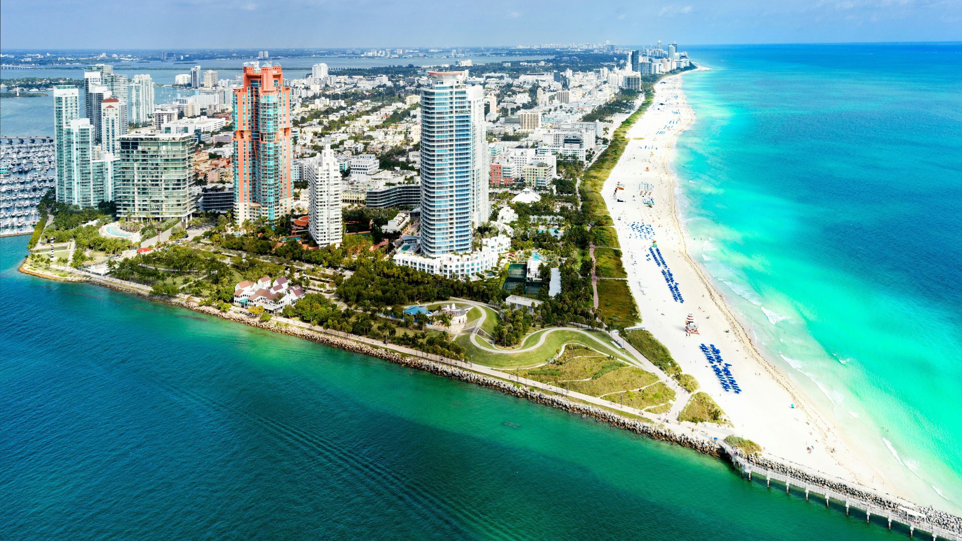 An aerial view of the stunning South Miami Beach in Florida, featuring a sandy beach with blue umbrellas, a pier, vibrant turquoise ocean water, and a backdrop of high-rise buildings and a cityscape under a blue sky.