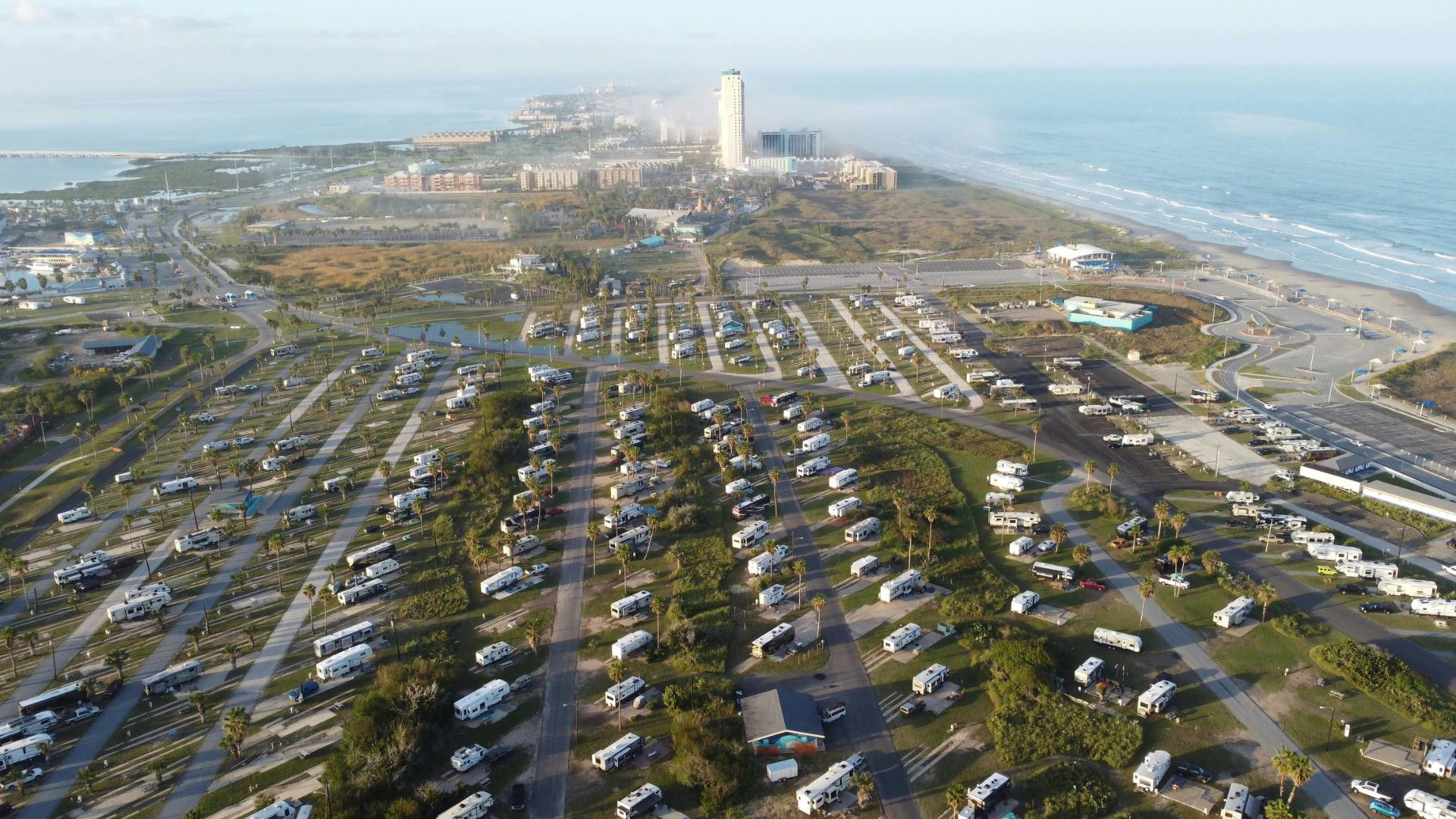 An aerial photograph showcasing a large, busy RV park packed with recreational vehicles and trailers near a sandy beach, with ocean waves breaking and a cluster of high-rise hotels in the distance under a bright, sunny sky.