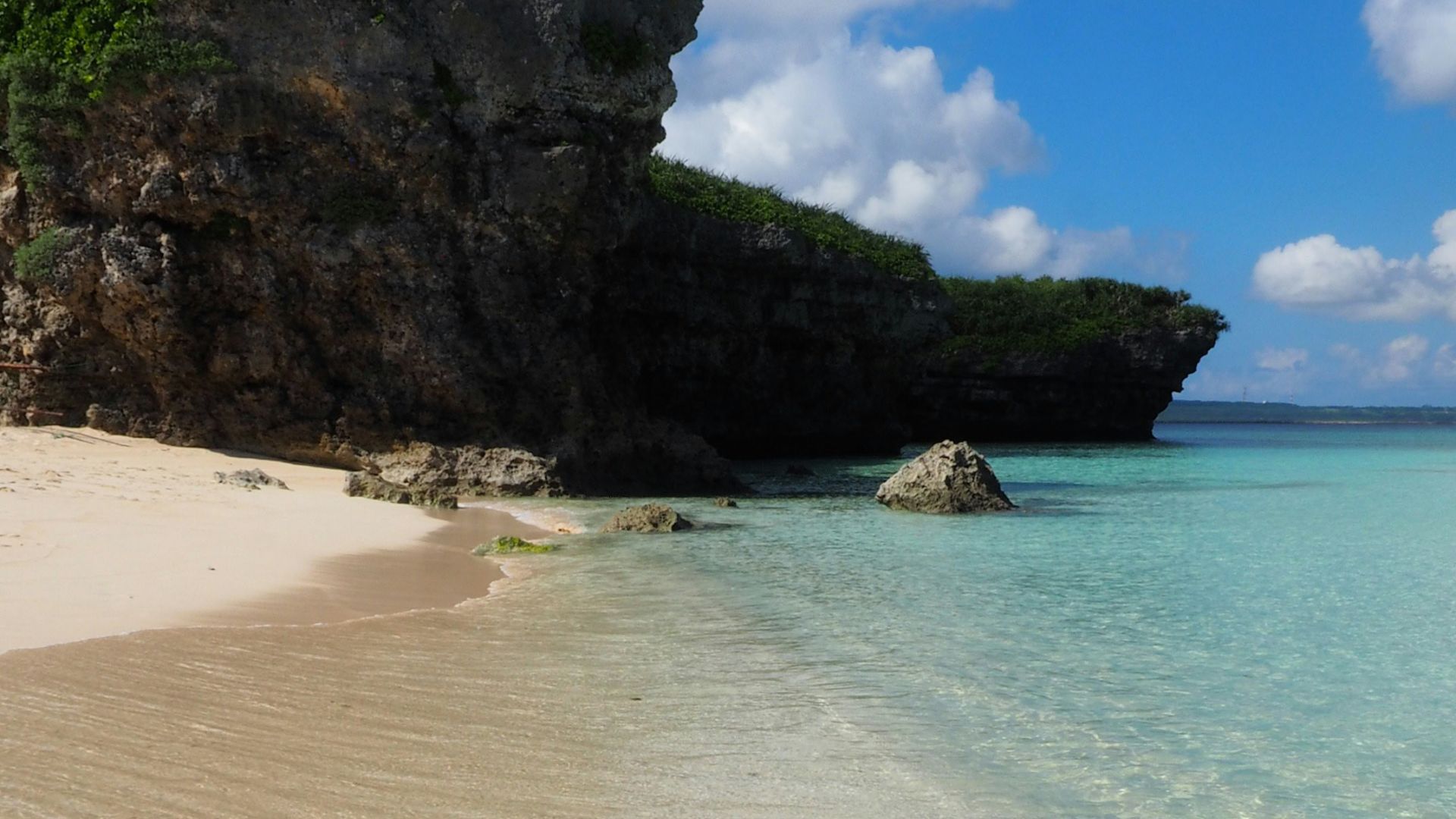A sandy white beach and exceptionally clear, shallow turquoise ocean water, with a large, dark, vegetated rock cliff formation rising dramatically on the left side under a bright blue sky with white clouds.