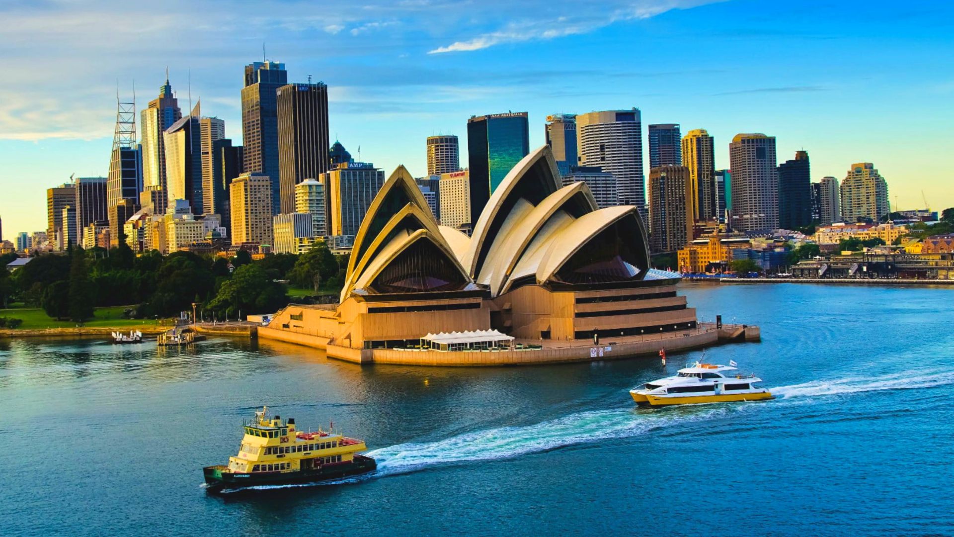 A bright, sunny daytime panoramic photograph of the Sydney, Australia skyline seen across a blue harbor, featuring the iconic spired Sydney Tower and the sail-shaped Opera House, framed by green trees in the foreground.