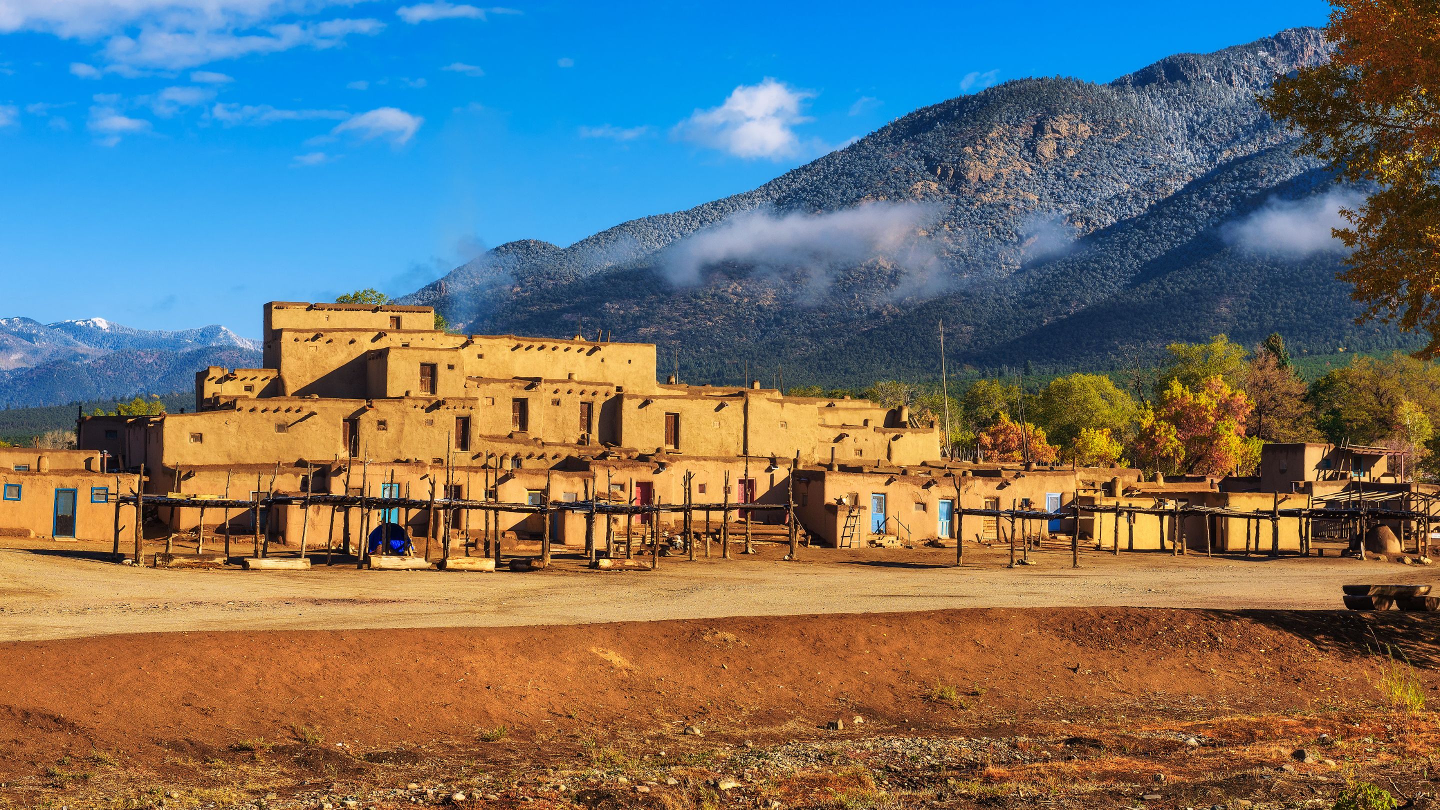 A photograph of the multi-storied reddish-brown adobe residential complex of the Taos Pueblo, an ancient Native American village in New Mexico, with wooden structures in the foreground and the large, rugged Sangre de Cristo Mountains behind it under a blue sky with white clouds.