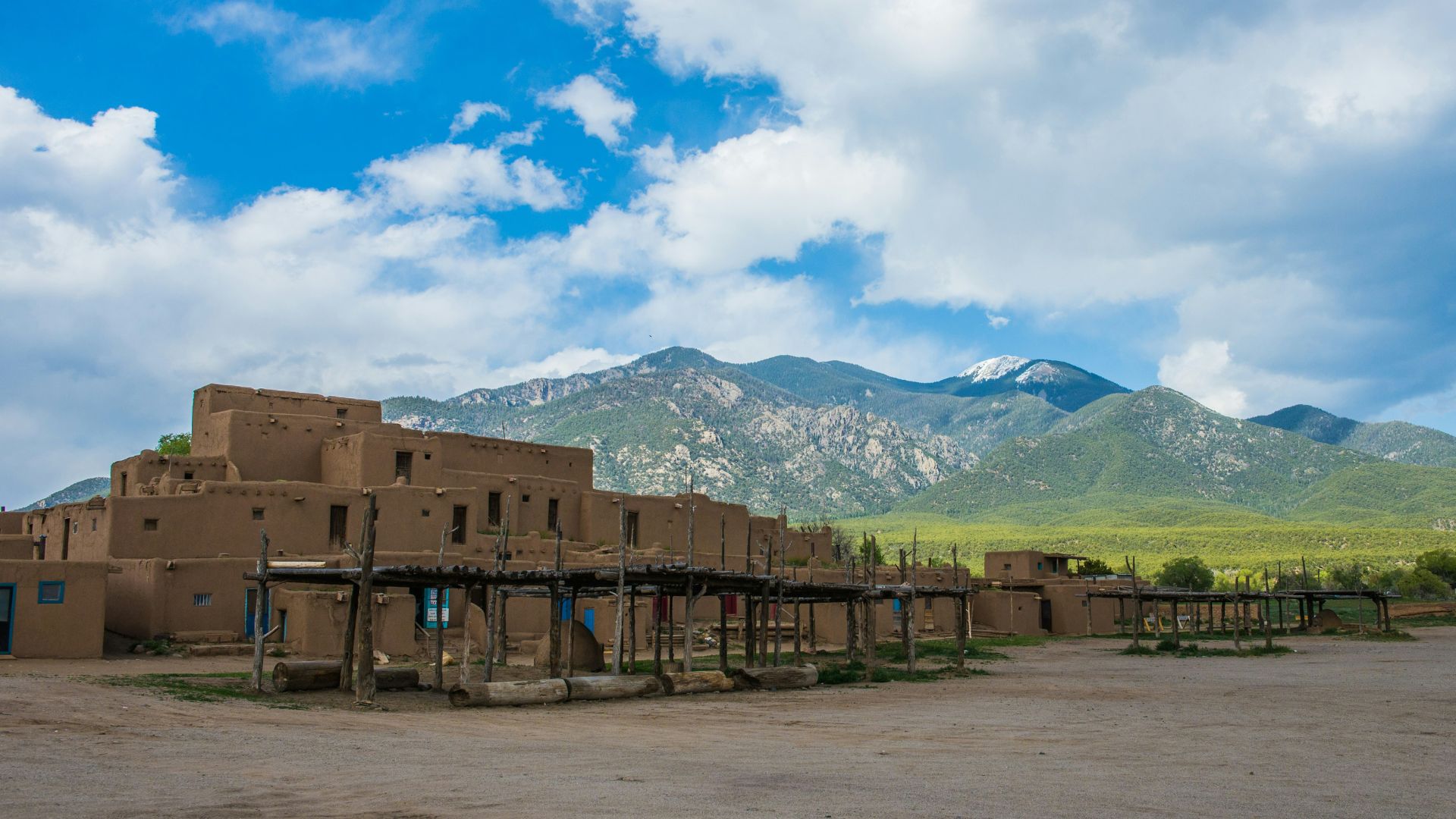 An iconic view of the historic, multi-storied reddish-brown adobe buildings of the Taos Pueblo Native American community in New Mexico, featuring exposed wooden beams and ladders, set against a backdrop of large green mountains under a bright blue sky with white clouds.
