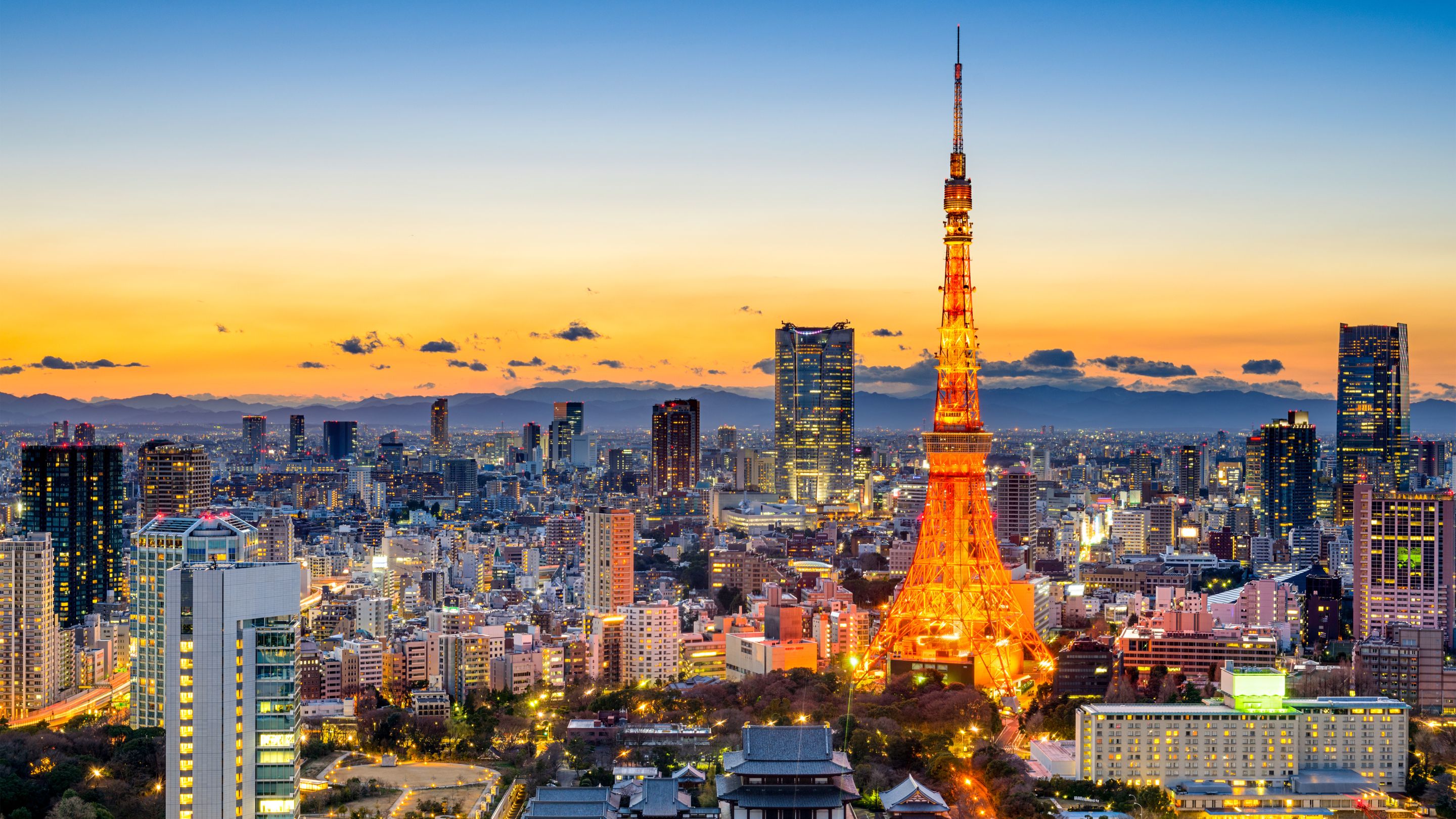 A panoramic night-time view of the brightly illuminated Tokyo Tower rising above a dense cityscape with numerous skyscrapers, set against a colorful yellow and orange twilight sky.