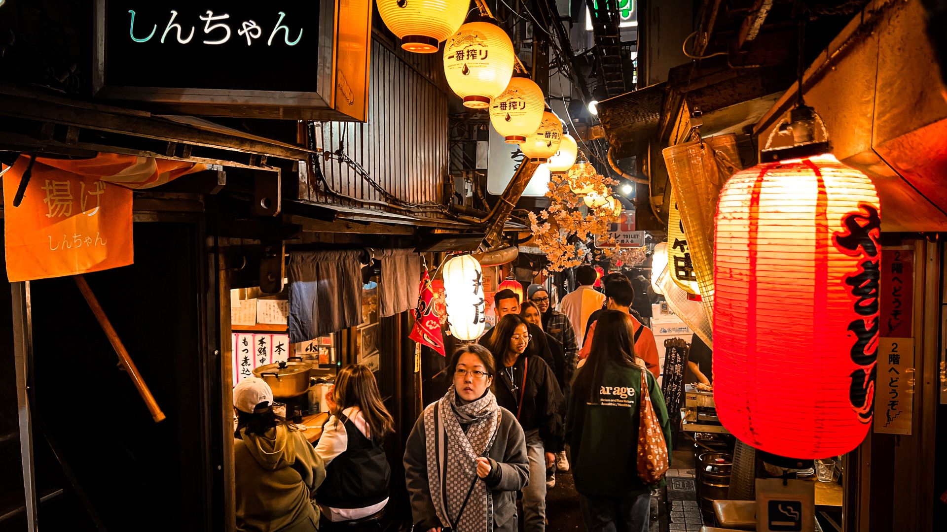 A crowded, narrow alleyway in Shinjuku, Tokyo, illuminated by numerous red and yellow paper lanterns hanging above small, traditional wooden izakaya bars and restaurants at night.