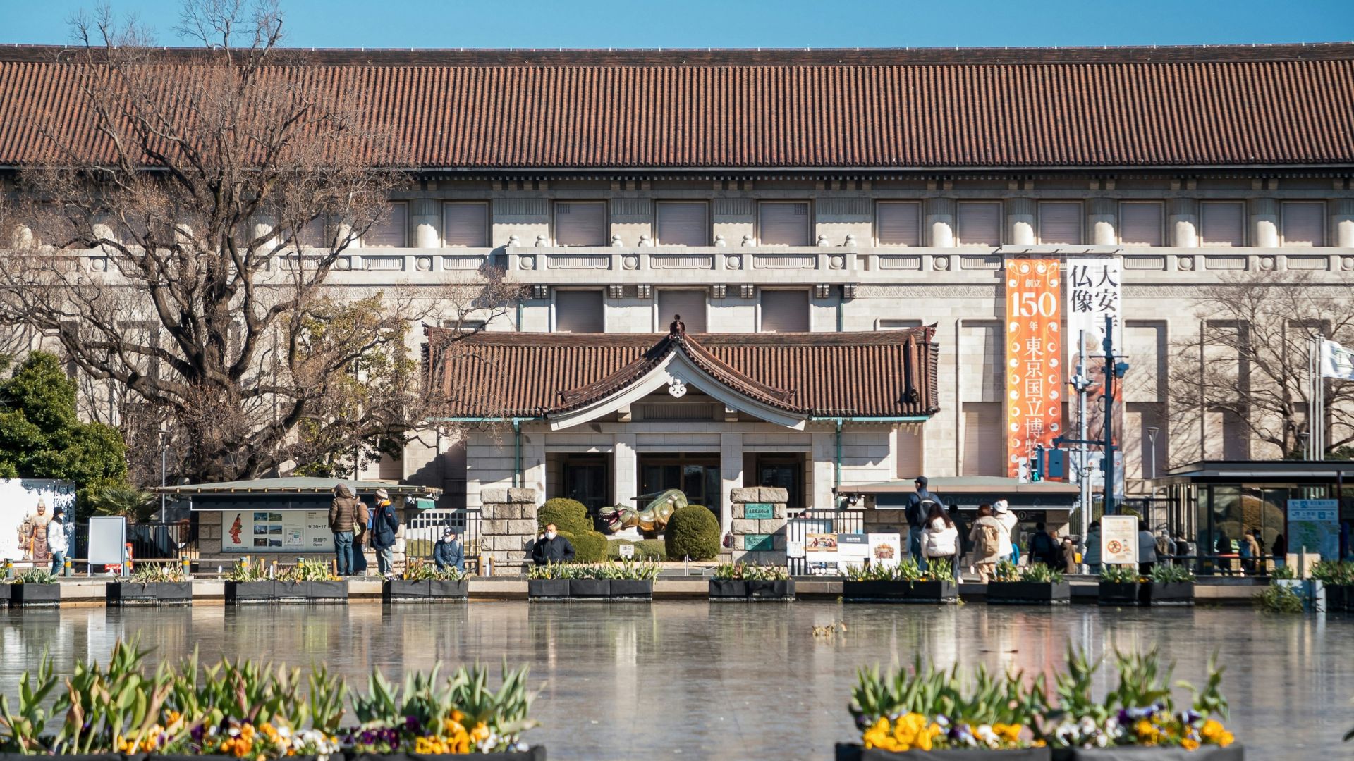 The large, traditional Japanese-style building of the Tokyo National Museum main gallery (Honkan) viewed across a calm reflecting pool with people milling about in the foreground.