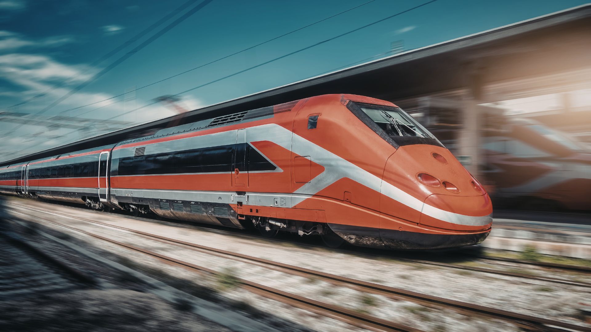 An orange and silver Trenitalia Frecciarossa 1000 high-speed train moving quickly along railway tracks outdoors under a blue sky.