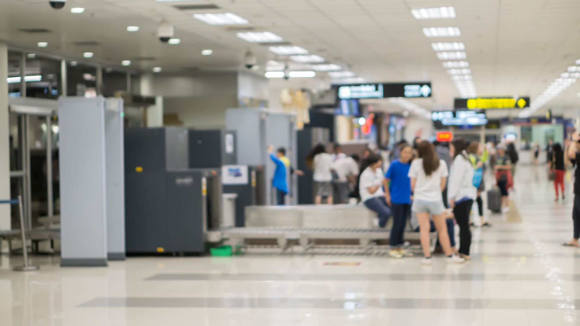 A blurred, generic stock photograph of an airport security checkpoint area with several passengers queuing for screening with their luggage.