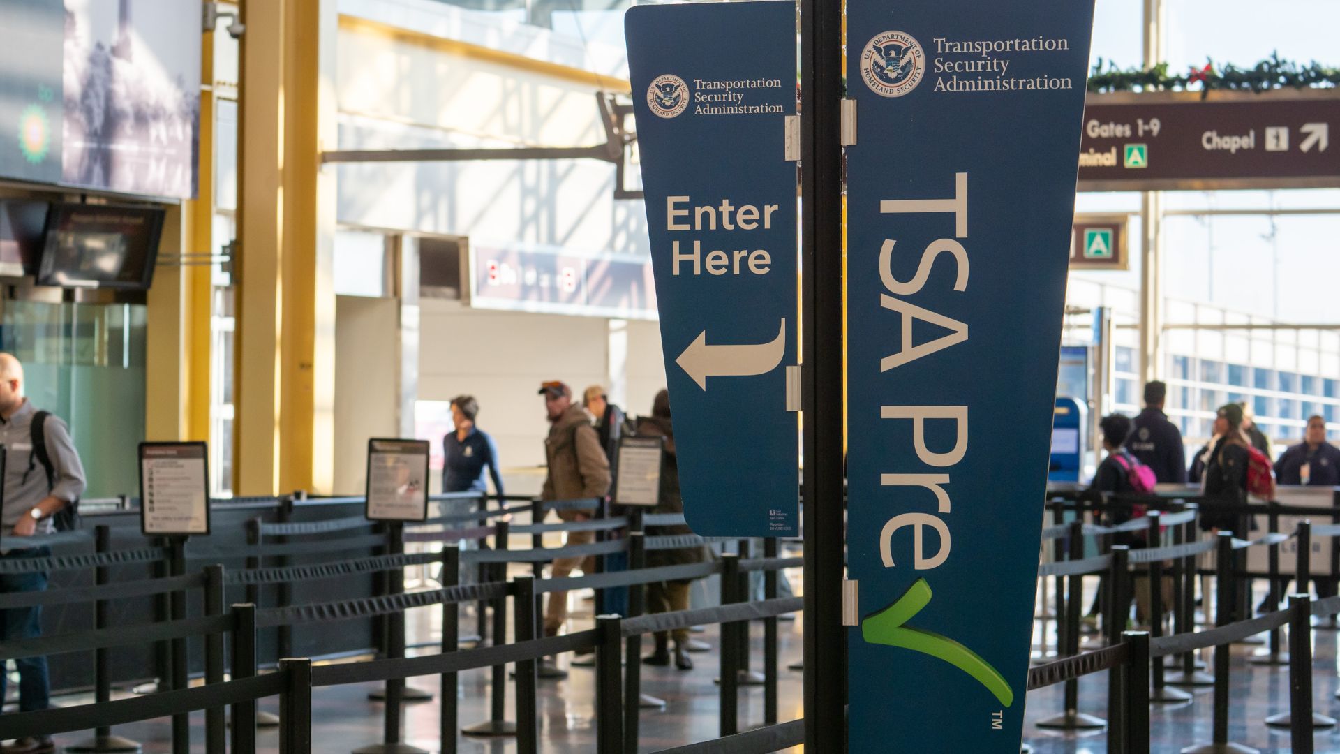 An interior photo of an airport security checkpoint at Ronald Reagan Washington National Airport (DCA), showing a busy queue area with prominent blue "TSA Pre✓®" and "Enter Here" signs.