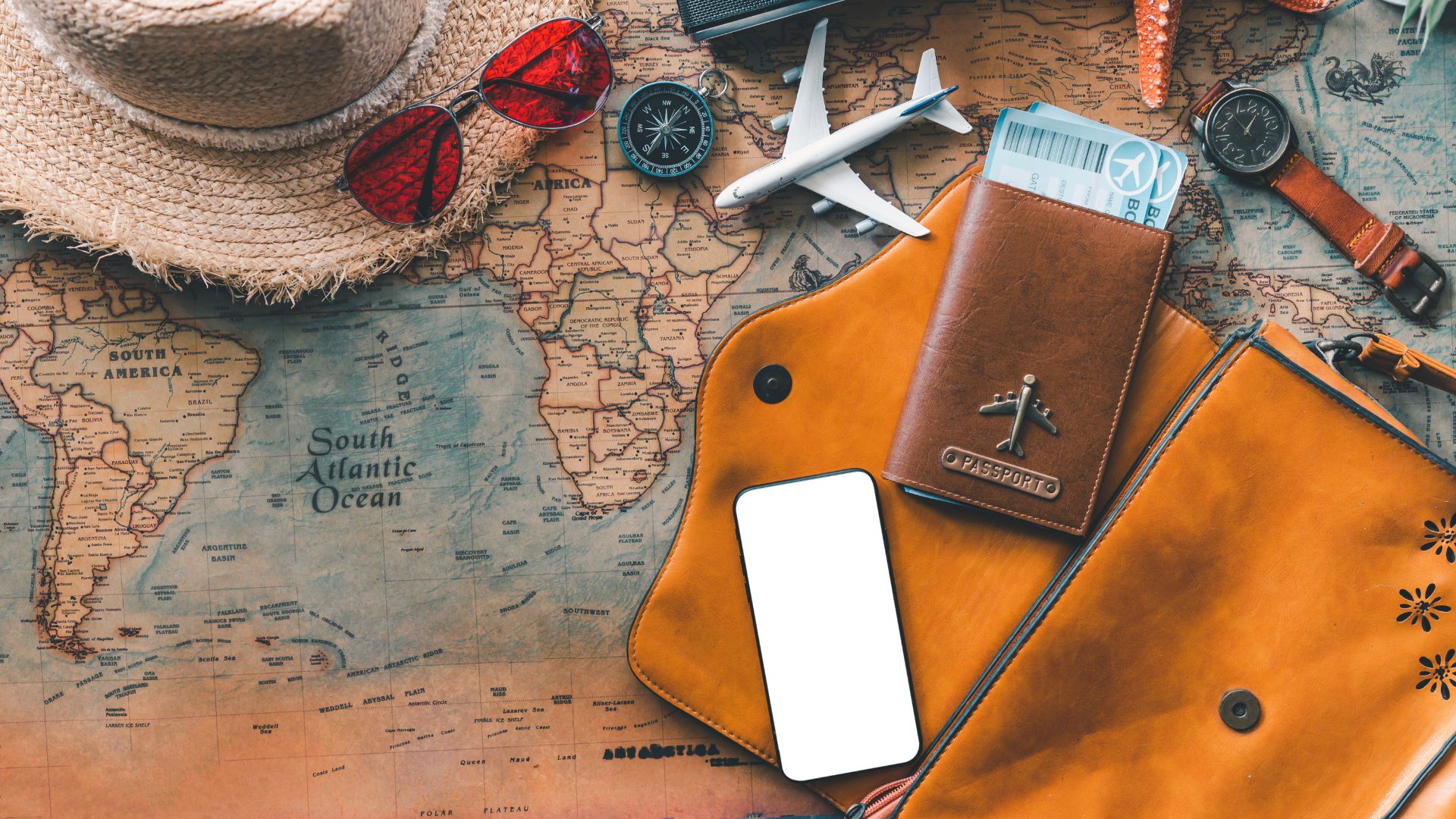 A flat lay arrangement of travel essentials, including a vintage map, a straw hat, red sunglasses, a passport in a leather holder, a model airplane, a compass, and a smartphone.