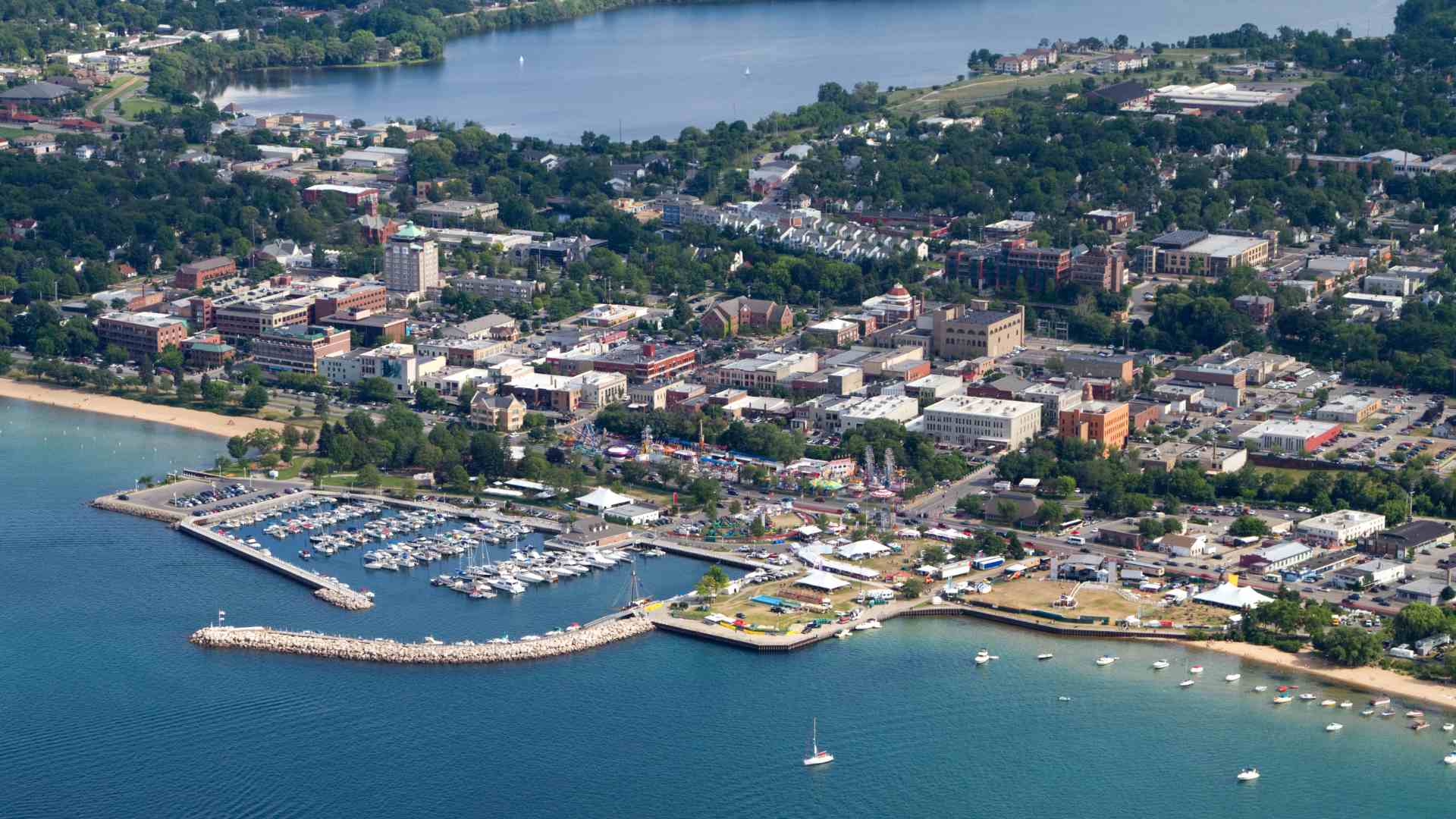 An aerial photograph of the downtown district of Traverse City, Michigan, showing a boat marina and park area on the large blue Grand Traverse Bay in the foreground, and a dense urban area extending inland to another smaller lake surrounded by green trees.
