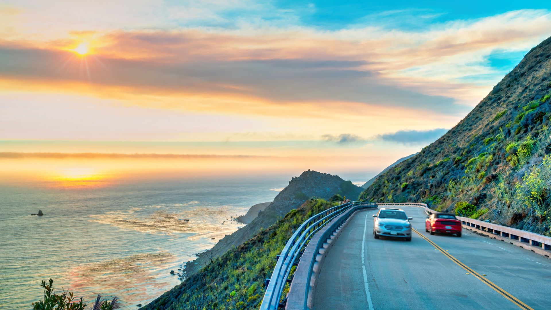 A breathtaking sunset photo of two cars driving along a winding, cliff-side section of the Pacific Coast Highway in Big Sur, California, overlooking the ocean.