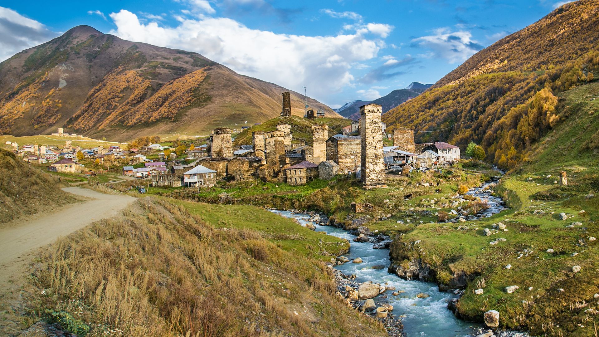 A scenic, daytime photo of the historic Ushguli village nestled in a lush green mountain valley in Georgia, featuring multiple ancient, square stone defensive towers (Svan towers) and a flowing river under a partly cloudy sky.