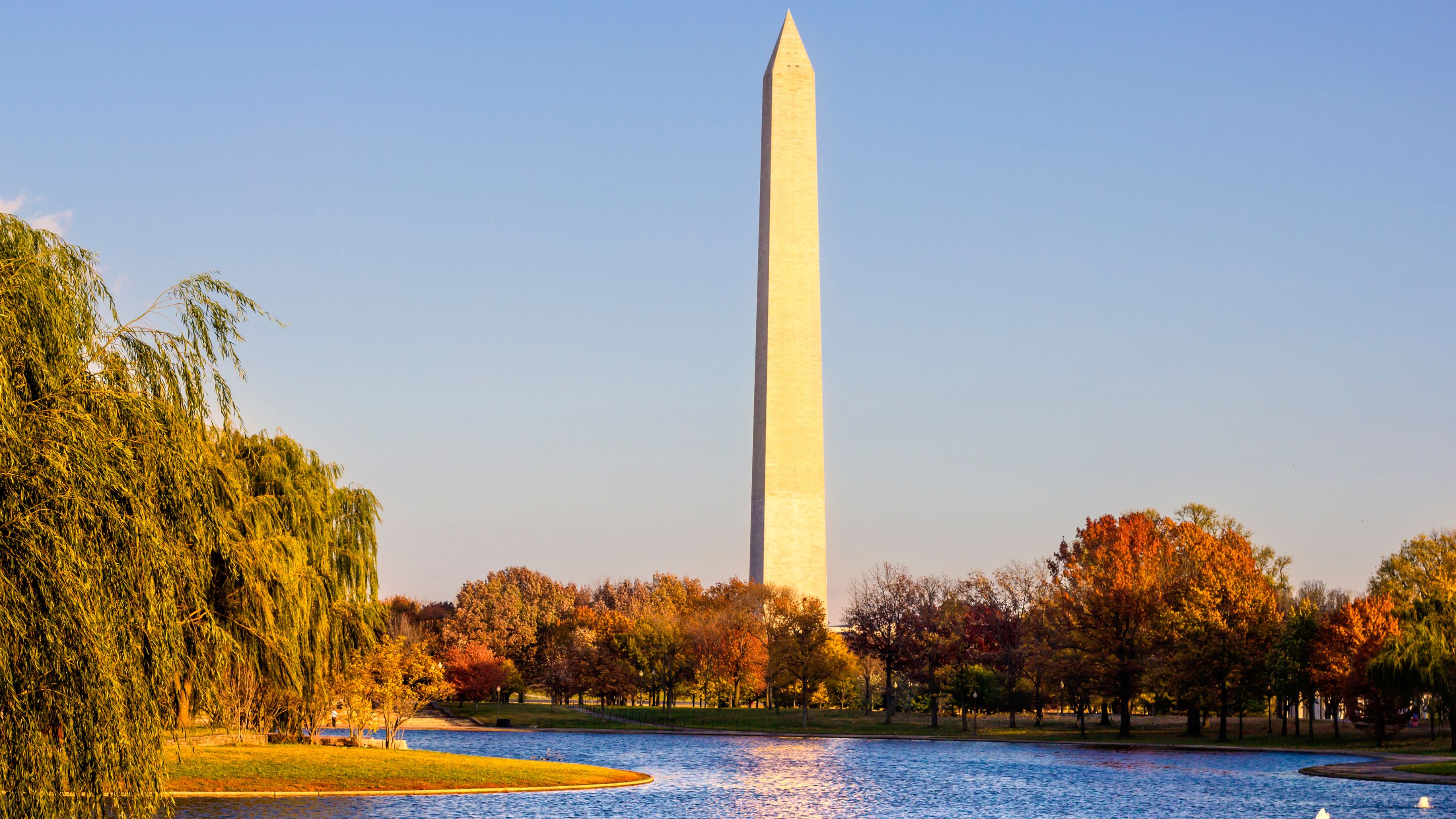 A tall, white obelisk known as the Washington Monument stands prominently in Washington, D.C., with a body of water and autumn-colored trees in the foreground under a clear, blue sky.