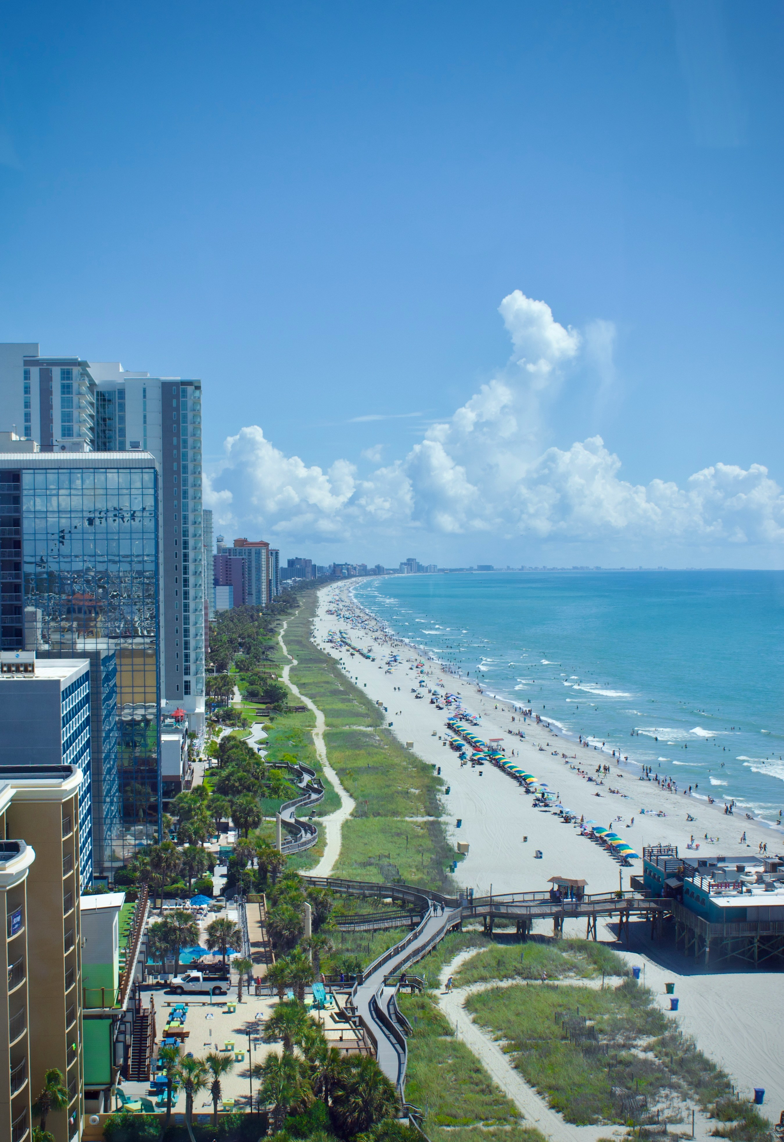 An aerial photograph looking down a long, wide, sandy beachfront lined with high-rise hotels and resorts, featuring a curved boardwalk system through beach grass and a pier extending into the bright blue Atlantic Ocean under a partially cloudy sky.
