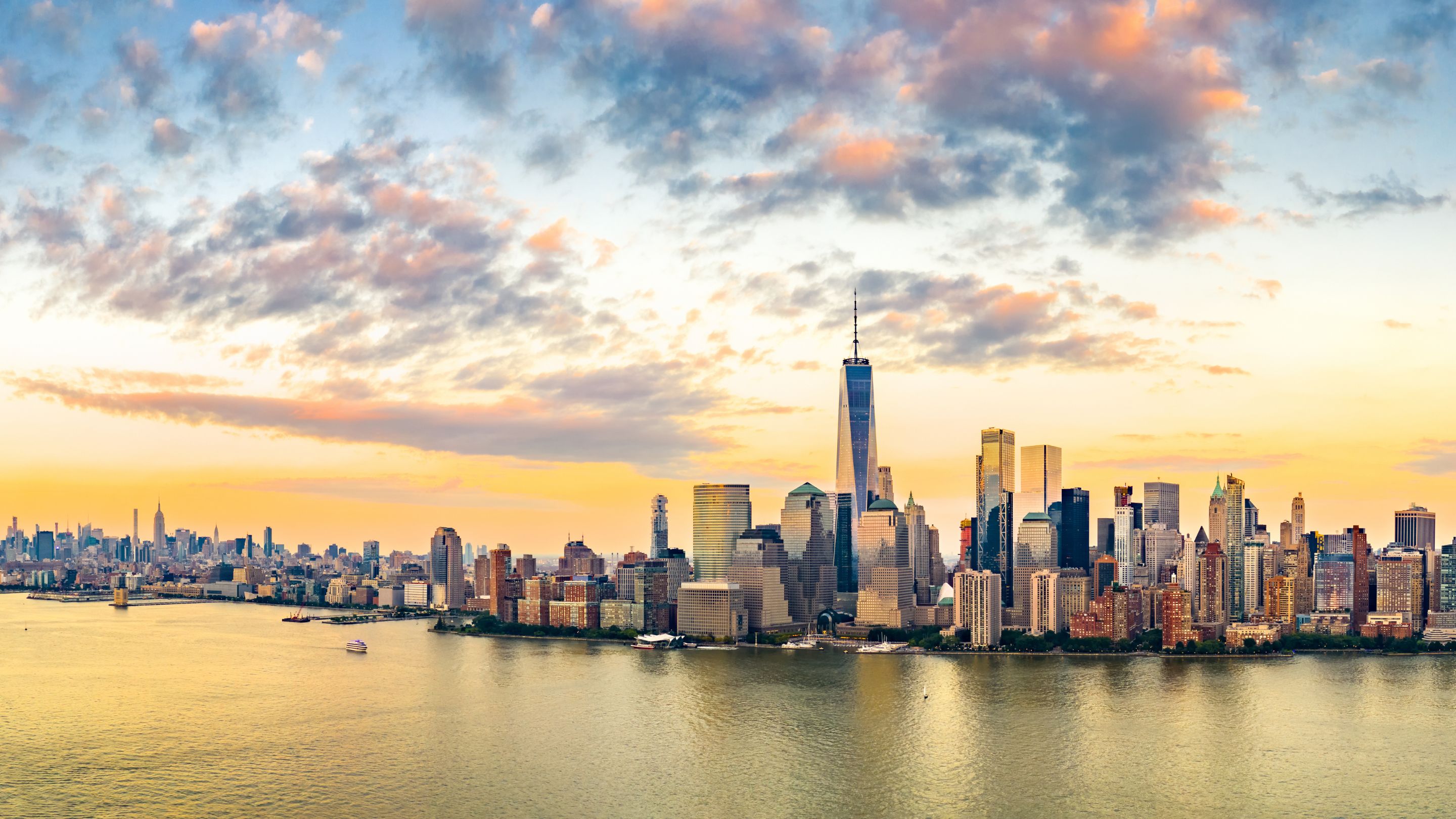 A panoramic view of the lower Manhattan skyline in New York City at sunrise or sunset, featuring the towering One World Trade Center skyscraper, other high-rise buildings, and the reflecting water of the Hudson River in the foreground.